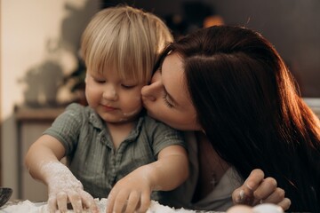 Tender kiss on flour-dusted cheek as toddler explores kitchen textures capturing authentic parenting slow family life and emotional connection in soft morning light during sensory cooking play