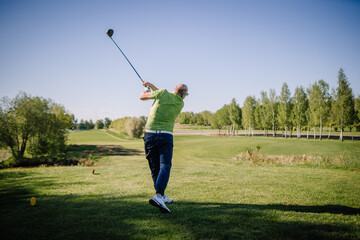 Man in green shirt completes a golf swing on a scenic course under clear blue sky, with trees and fairway stretching into the distance..