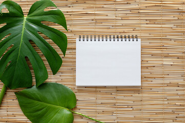 Top view flatlay of a blank white page of spirallled notebook on straw reed shade suface with two green monstera leaves
