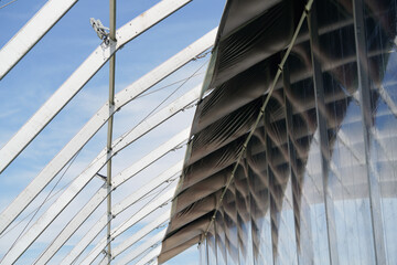 A close-up view of a modern architectural structure with a curved, glass-covered roof supported by white beams. The sky is visible through the structure's framework.