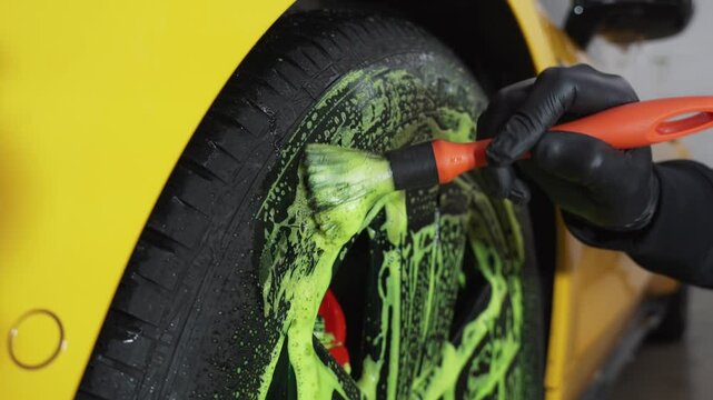 Detailing procedure for car care at the service 4k video. Cleaner washes car tire with green foam using brush with detergent at the car wash.