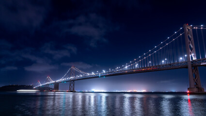 Night view of the Bay Bridge with lights reflecting on the water.