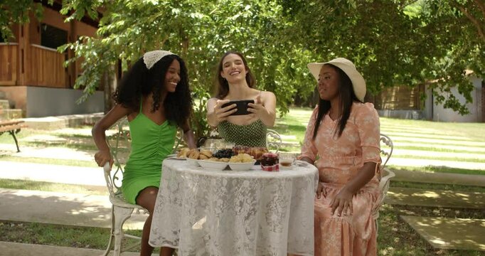 Diverse female friends taking selfie sitting at table at outdoor brunch.