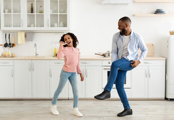 Kitchen Fun. Full length of joyful black dad and his little daughter singing and dancing while cooking together at kitchen, girl using whisk as microphone, father playing on leg as on guitar