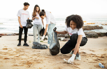 Happy young multiethnic people volunteers in t-shirts with garbage packages clean up garbage, plastic bottles on sea beach, outdoor. Environment conservation, protecting eco