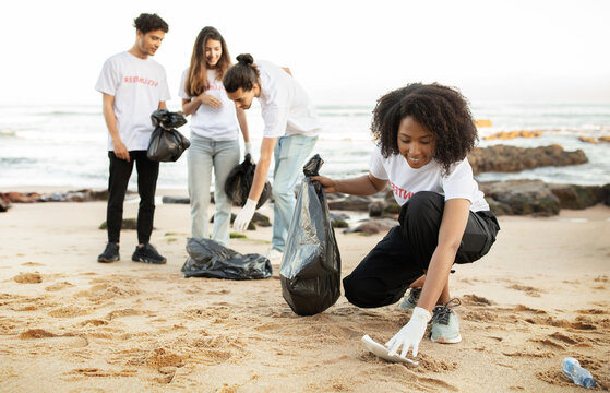 Happy young multiethnic people volunteers in t-shirts with garbage packages clean up garbage, plastic bottles on sea beach, outdoor. Environment conservation, protecting eco