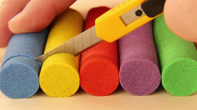 Colorful kinetic sand cutting ASMR. Yellow plastic knife slices through blue, yellow, red, purple and green sand cylinders creating satisfying effect.
