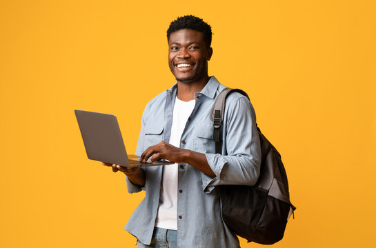 Cheerful african american male student positive guy in casual outfit with backpack using modern laptop on yellow studio background, smiling at camera, enjoying nice educational website, copy space