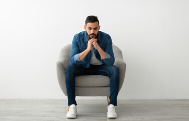 Negative emotions. Young Arab man sitting in armchair with dull or thoughtful face expression,...