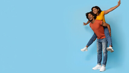 Cheerful black guy giving piggyback ride to his happy pretty girlfriend while posing over blue...