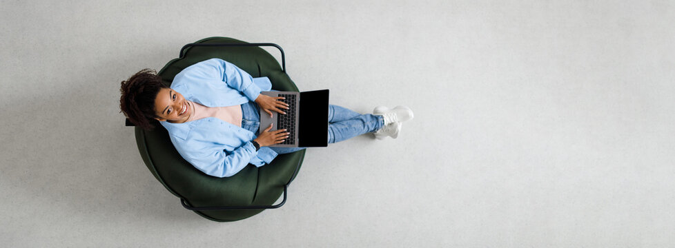 People And Modern Technology. Above top view of young smiling black woman holding pc on lap sitting on bean bag in living room, looking up at camera. Lady browsing internet, free copy space panorama