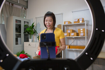 An Asian woman in a yellow t-shirt is live streaming as she sells clothes, engaging warmly with...