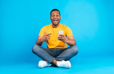 Excited African American Man Holding Smartphone, Pointing Finger At His Mobile Phone In Excitement Sitting Over Blue Background In Studio. Joyful Guy Recommending Application For Your Cellphone