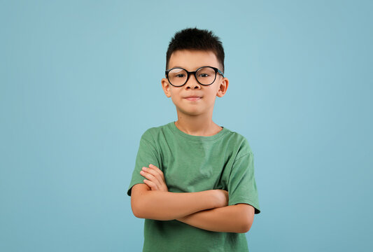 Portrait of smiling little asian boy wearing eyeglasses standing with folded arms and looking at camera, confident nerdy korean male child posing over blue studio background, copy space