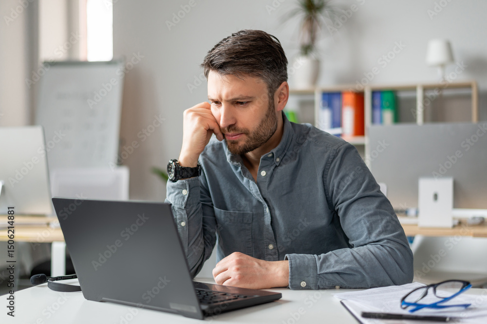 Poster serious middle aged businessman using laptop computer and reading reports, sitting at workplace in o