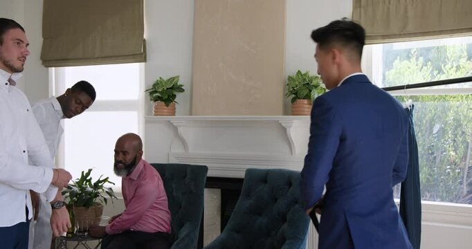 After sipping drink, diverse male friends tying navy necktie and fixing cufflinks on groom at home