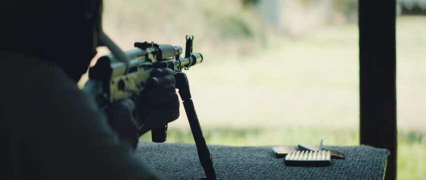 Man lies prone and fires with rifle on bipod aiming at distant target in open shooting range during outdoor training session