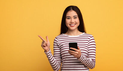 A cheerful young Asian woman holds a smartphone in one hand while pointing with the other. She wears a striped shirt, and the bright yellow background enhances her joyful demeanor.