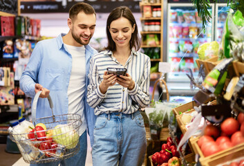 Smiling Millennial Couple Shopping Groceries In The Hypermarket, Two Happy Consumers Buying Food Walking With Cart In Wholesale Shop, Choosing Eco Vegetables And Fruits, Checking Label And Expiry Date
