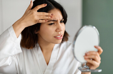 Upset arabic young woman in white silky robe looking at hand mirror, checking her skin, touching...
