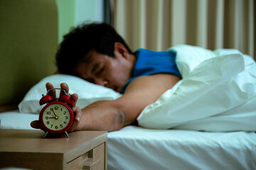 Sleeping man in bed reaches for ringing alarm clock, representing early morning wake-up or reluctance to get up