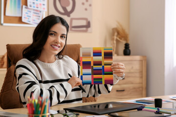 Young female graphic designer working with tablet and color palettes at table in office