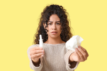 Young African-American woman with handkerchief and nasal spray suffering from allergy on yellow background