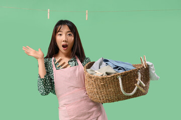 Shocked young Asian woman with laundry basket and hanging clothespins on green background