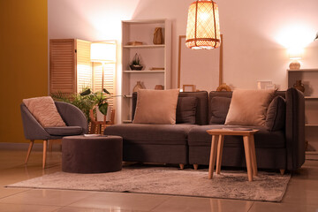 Interior of modern living room with grey sofa, armchair, shelving unit and glowing lamps at evening