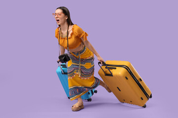 Young female tourist carrying heavy suitcases on lilac background