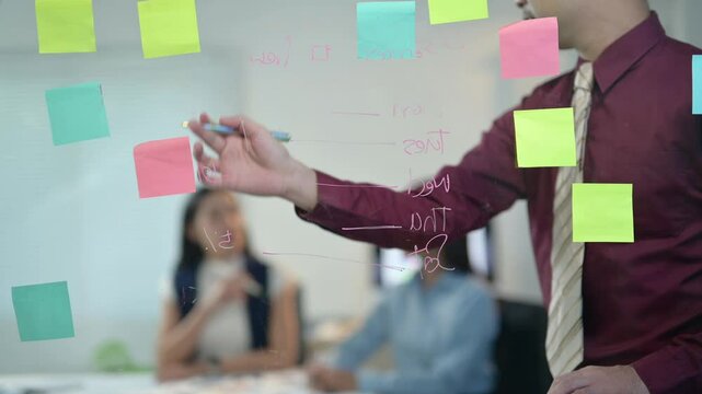 A man is writing on a whiteboard with colorful sticky notes. The board has a lot of notes on it, and the man is pointing to one of them. Scene is focused and productive