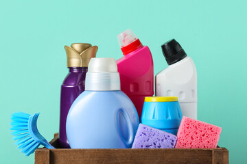 Box with cleaning supplies and bottles of detergent on turquoise background, closeup
