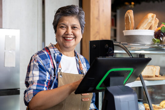 Asian senior woman with short gray hair wearing plaid shirt and brown apron smiling behind counter using touchscreen at cashier station inside small family-owned bakery shop - Powered by Adobe