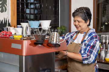 Fototapeta premium Asian senior woman in plaid shirt and apron smiling while using espresso machine making coffee at cozy cafe interior with colorful cups and brewing tools on counter in warm atmosphere