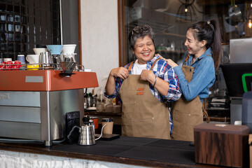 Asian senior woman smiling joyfully while young Asian female coworker helping adjust apron in warm...