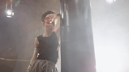 A woman in a boxing ring, tiredly leaned on a punching bag to regain her breath after an intense workout. The photo is in a cinematic style, the light gives the mood to a sports motivational story