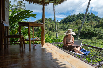 Sitting in a hammock on the terrace of a bamboo hut, a woman dons a straw hat while working on her laptop amid the vibrant jungle. Sunlight filters through the leaves, creating a serene atmosphere. 