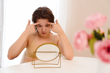 Stressed sad brunette young woman wearing beige top looking at mirror and touching her face having...