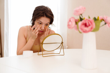 Stressed sad brunette young woman wearing beige top looking at mirror and touching her face having...