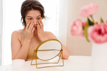 Stressed sad brunette young woman wearing beige top looking at mirror and touching her face having...