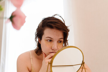 Stressed sad brunette young woman wearing beige top looking at mirror and touching her face having...