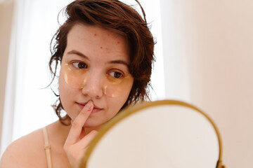 Stressed sad brunette young woman wearing beige top looking at mirror and touching her face having...