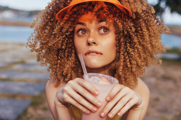Young woman with curly hair enjoying a refreshing drink outdoors in a vibrant setting