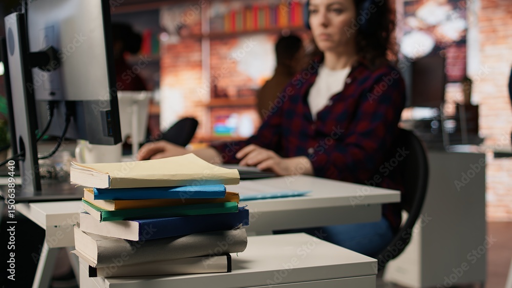 Poster focus on stack of books on startup office desk with woman in blurred background typing on pc, solvin