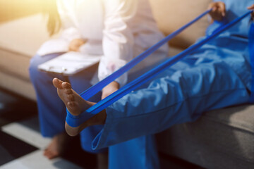 Fototapeta premium Confident physical therapist helps patient use resistance band stretching out his leg in clinic room.