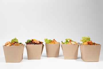 Five open eco-friendly brown paper boxes filled with various delicious and colorful Asian-style noodle and rice dishes, ready for takeaway , set against a clean white background.