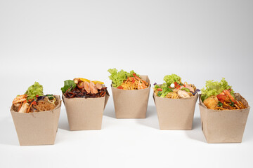 Five open eco-friendly brown paper boxes filled with various delicious and colorful Asian-style noodle and rice dishes, ready for takeaway , set against a clean white background.