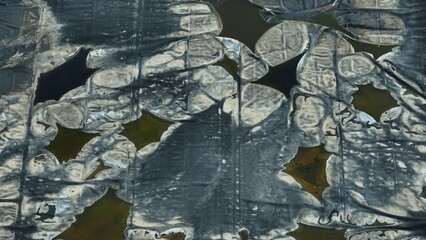 Aerial view of cracked geomembrane linings over waste containment ponds. Irregular patches of liquid indicate seepage or residue accumulation, highlighting degradation in the containment structure.
