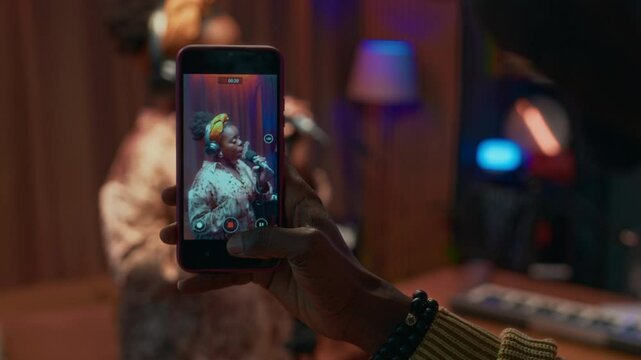 Handheld shot of young African American man filming female signer on smartphone in vibrant recording studio