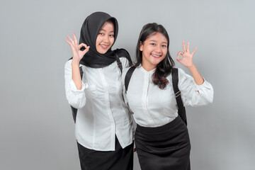 Two happy Asian high school girls in uniform, smiling and showing OK hand signs, ready to go back to school. Studio shot with grey background, education concept.